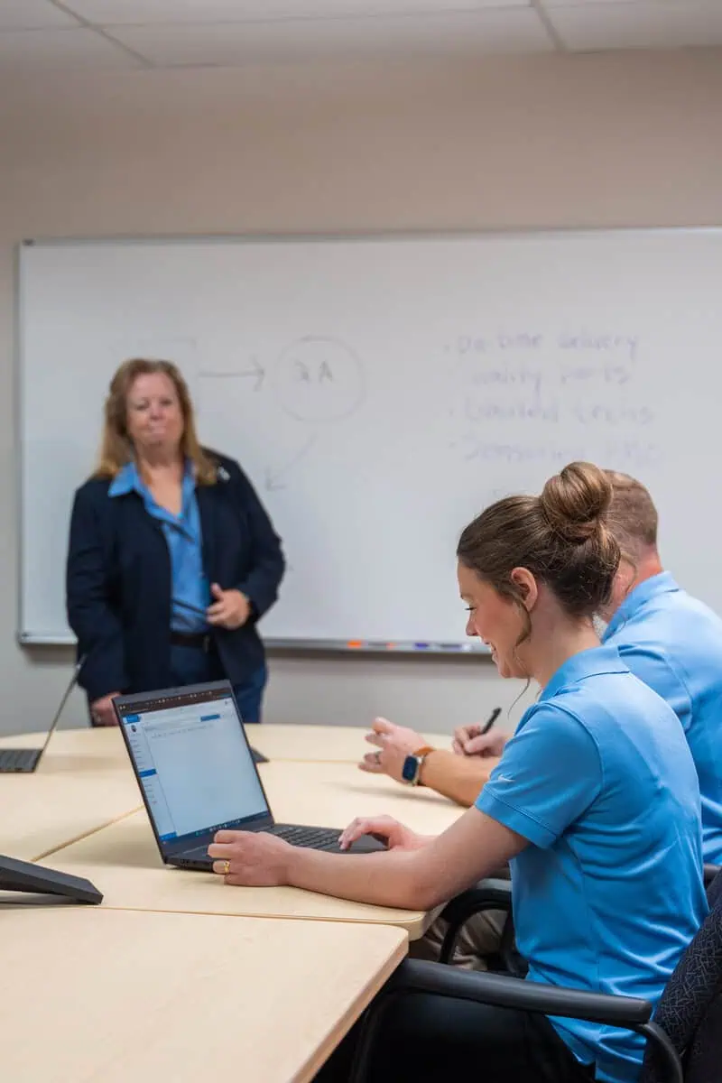 Technician training session with a woman presenting at a whiteboard, attendees using laptops for tech support.