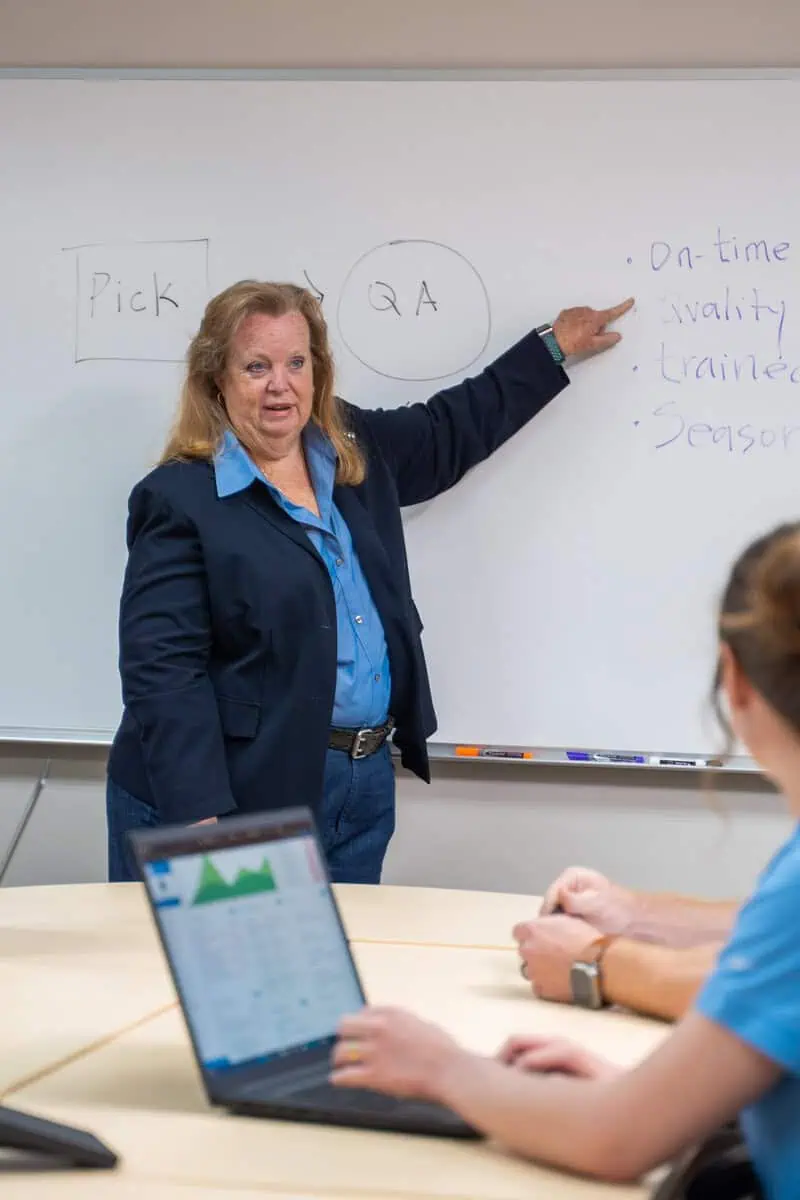 Woman leading a meeting on warranty management, pointing to a whiteboard with notes on quality and timelines.